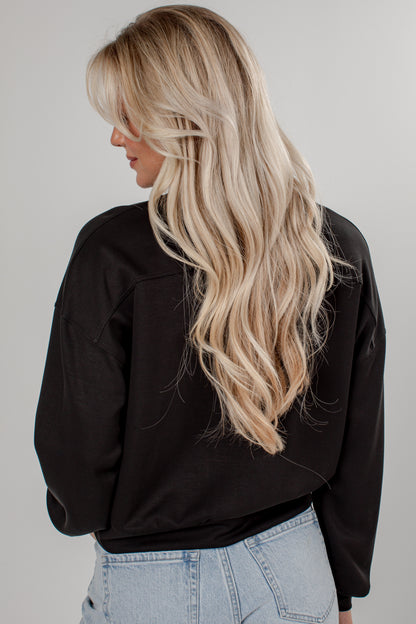 A woman with long, wavy blonde hair is shown from the back, wearing a black long-sleeve top and light blue jeans against a plain light gray background.