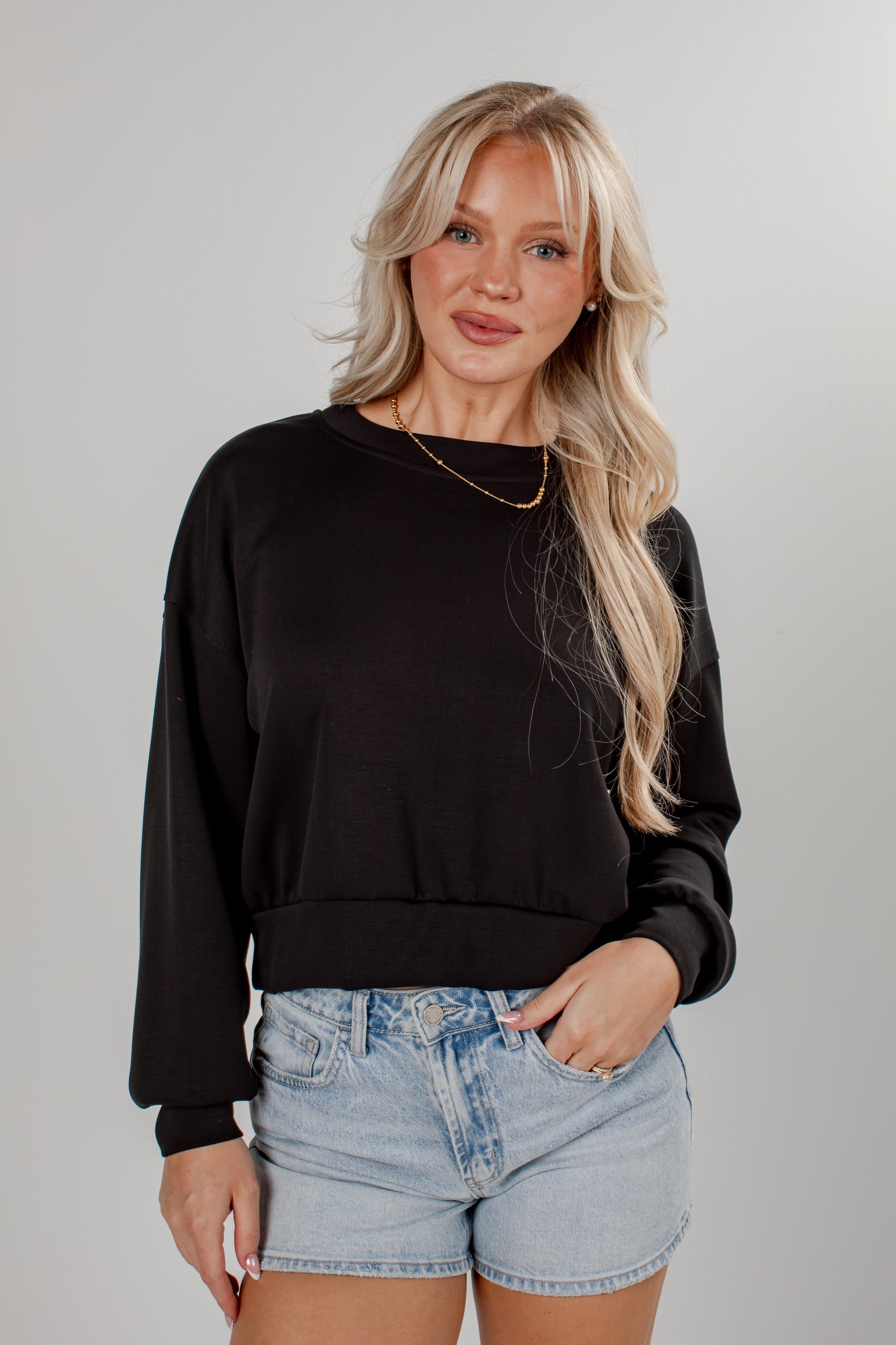 A blonde woman wearing a black long-sleeve crewneck sweatshirt and light-wash denim shorts, standing against a neutral gray studio background.