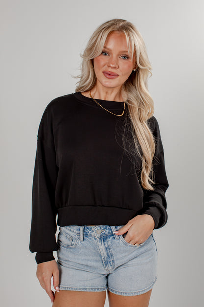 A blonde woman wearing a black long-sleeve crewneck sweatshirt and light-wash denim shorts, standing against a neutral gray studio background.