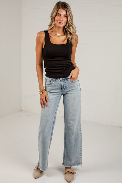 A woman modeling a black ribbed tank top and light-wash wide-leg denim jeans with frayed hems, standing against a white studio wall.