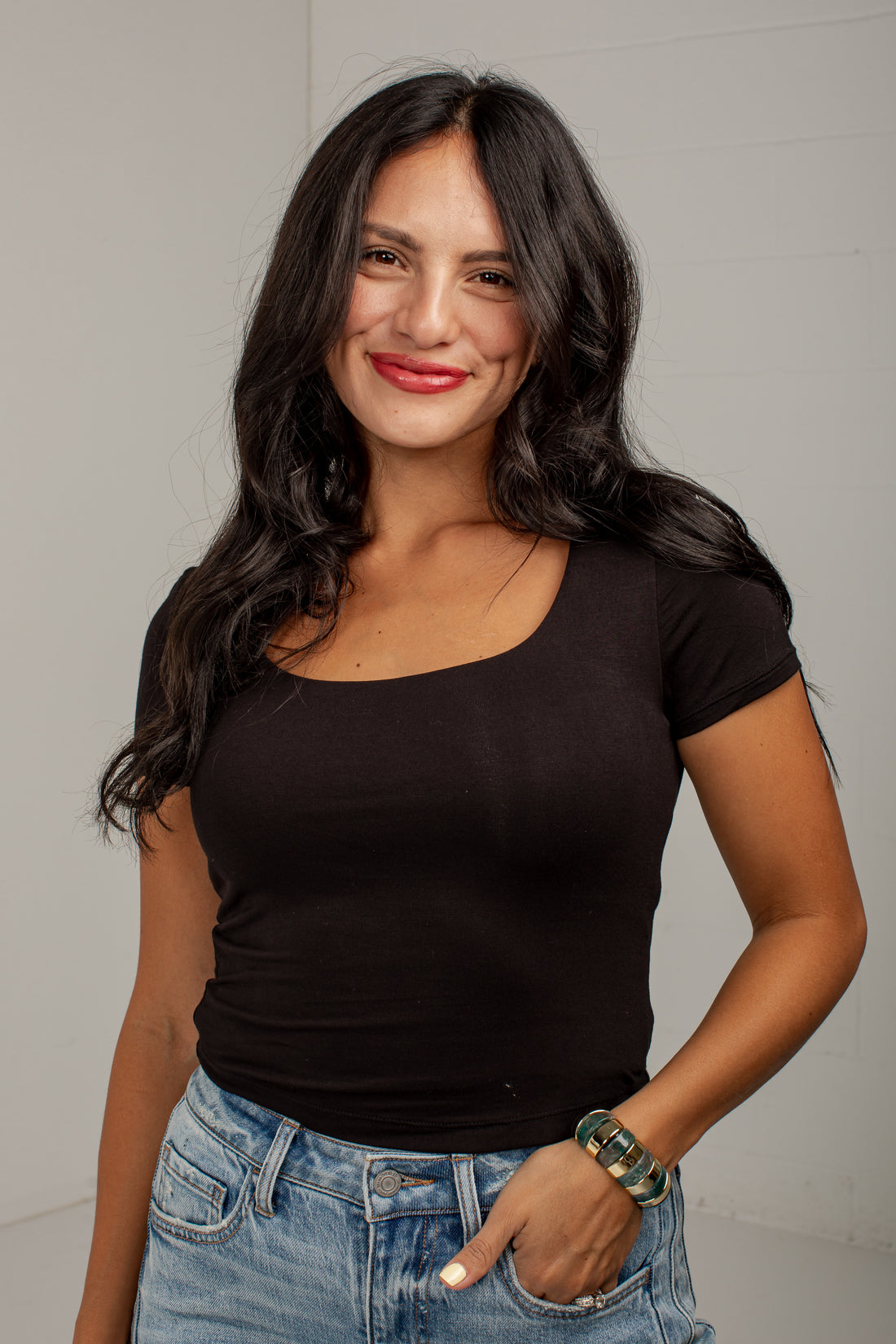 A woman with long dark hair wearing a fitted black scoop-neck short-sleeve top and light-wash blue jeans, standing against a light gray wall.