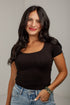 A woman with long dark hair wearing a fitted black scoop-neck short-sleeve top and light-wash blue jeans, standing against a light gray wall.