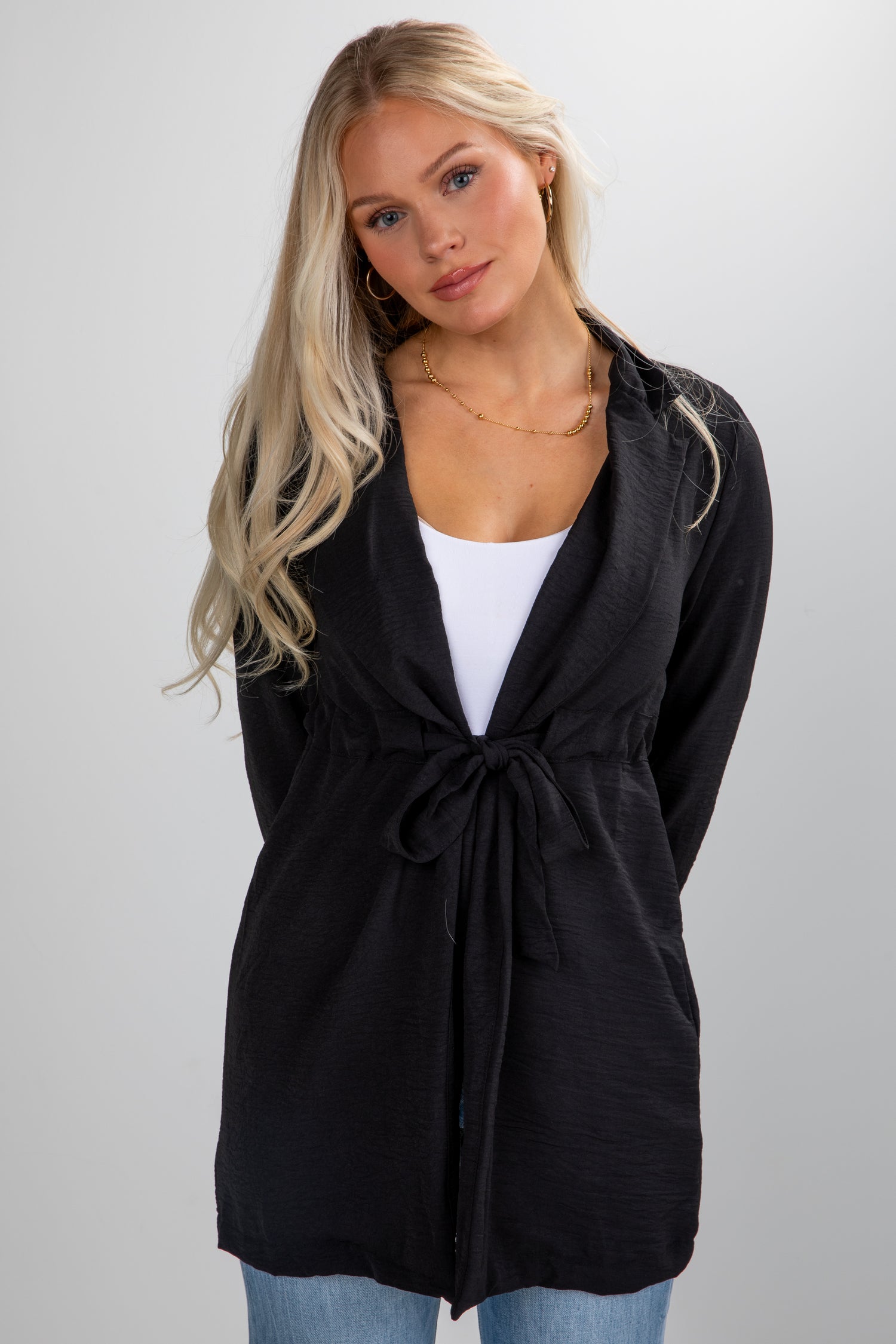 Woman wearing a black cardigan over a white top and blue jeans on a plain background