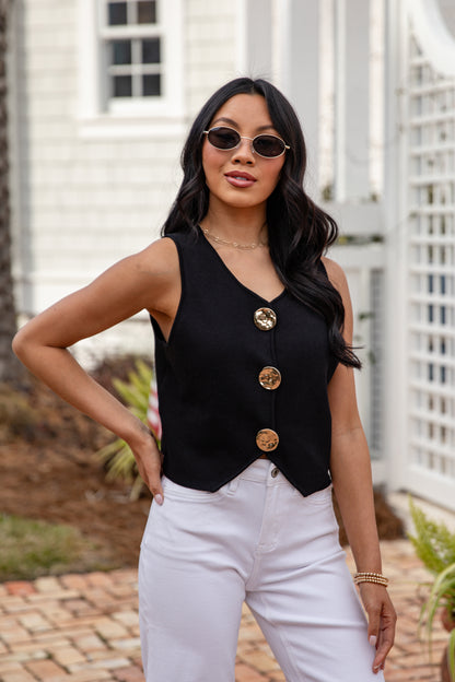 Woman wearing a black sleeveless top with gold buttons and white pants outdoors.