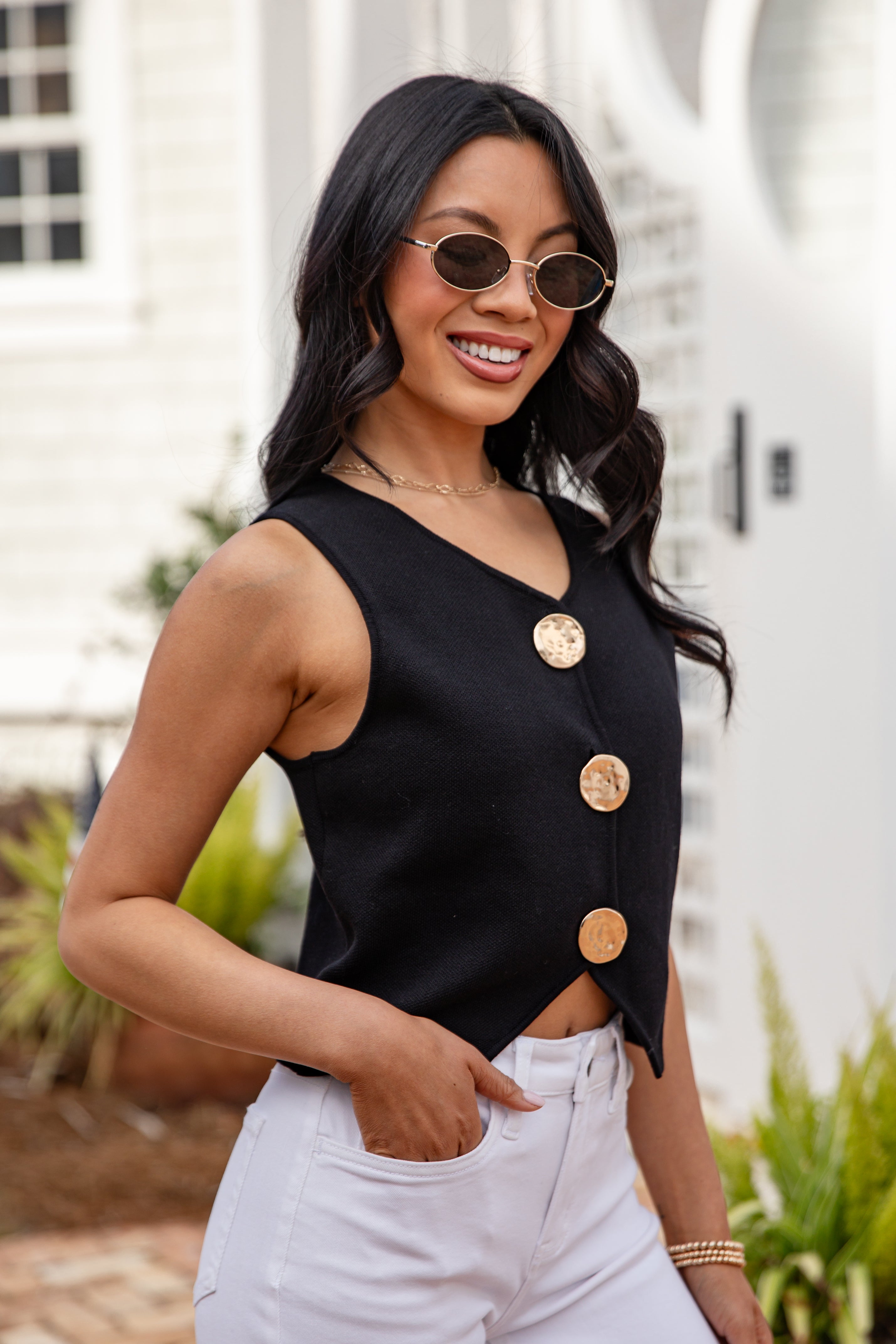 Woman wearing a black sleeveless top with gold buttons and white pants outdoors.