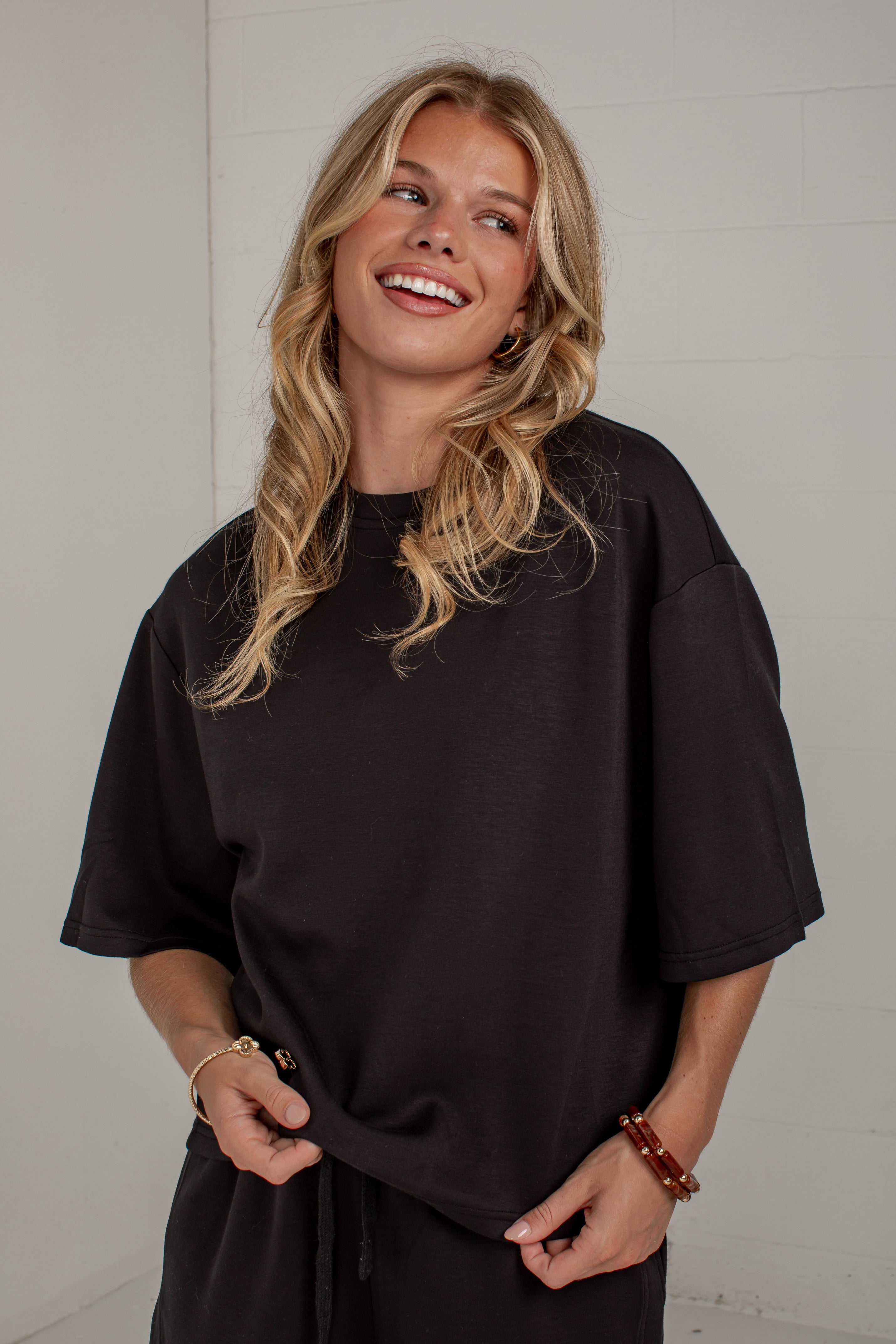 A smiling blonde woman wearing an oversized black short-sleeve t-shirt and matching black pants, standing against a white paneled wall.