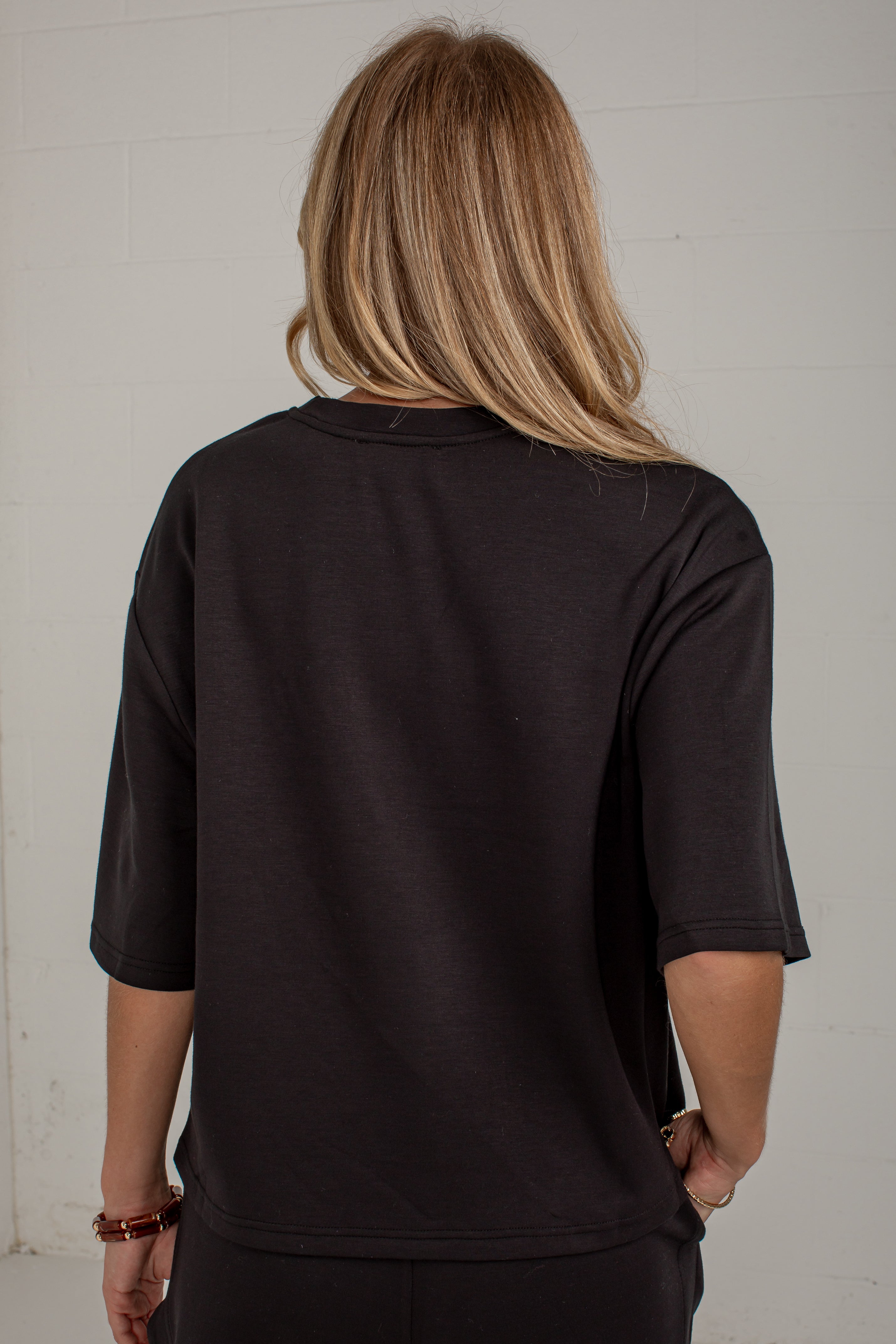 A woman viewed from behind wearing a black short-sleeve t-shirt and black pants, standing against a white brick wall.