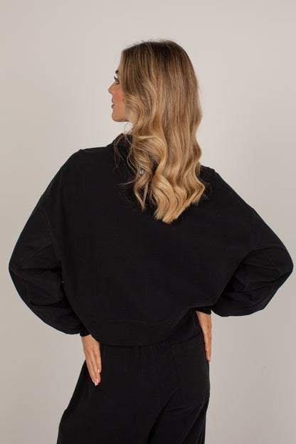 A woman with blonde wavy hair is shown from the back wearing a black oversized long-sleeve top and black pants against a plain light gray studio background.