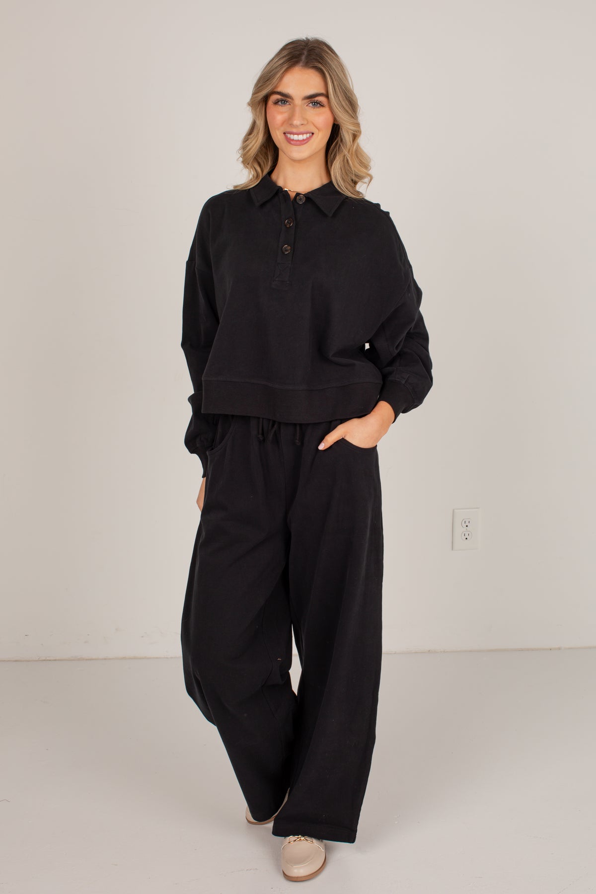A woman modeling a black long-sleeve collared button-front top with matching black wide-leg drawstring pants, standing against a plain light gray studio wall.