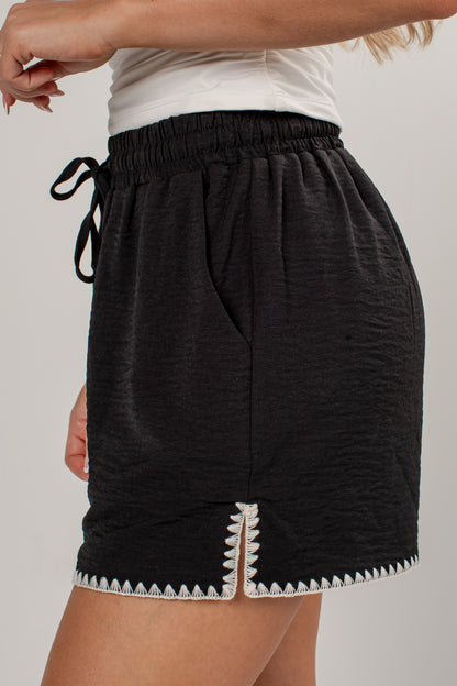 Black textured mini shorts with a white geometric patterned trim along the hem and a black drawstring waist, shown on a model.