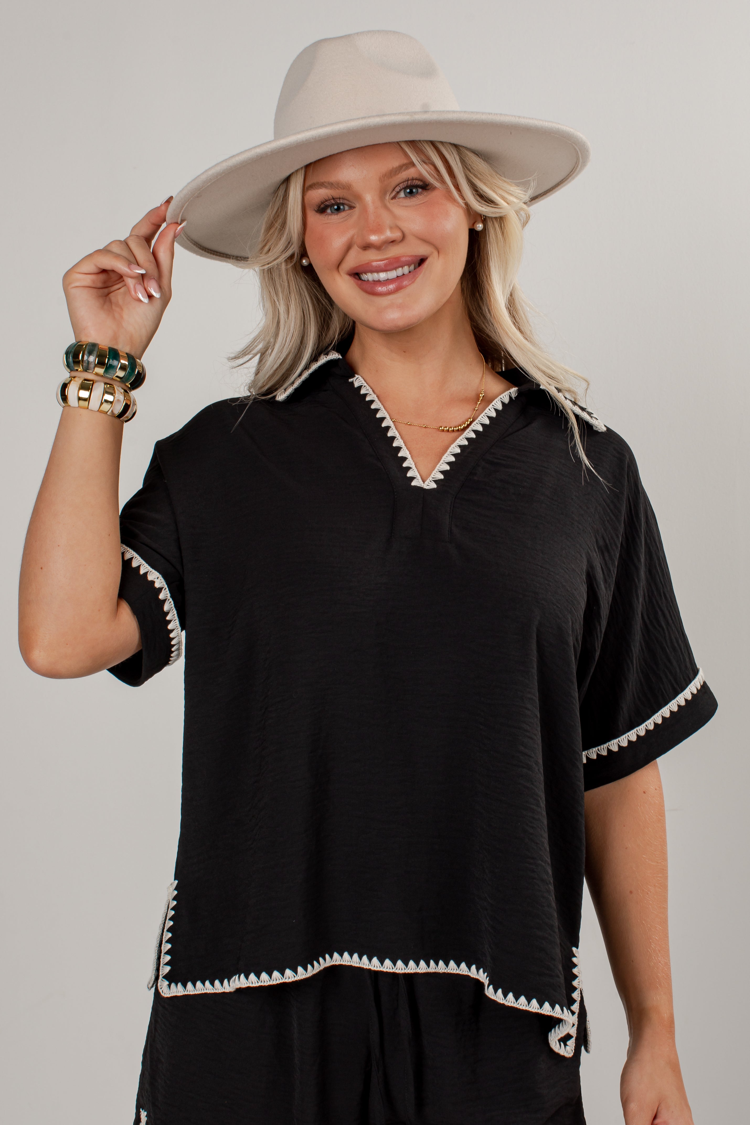 A woman wearing a black short-sleeve top with white geometric trim, a beige wide-brim hat, and gold bracelets, posing against a light gray studio background.