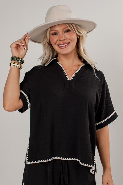 A woman wearing a black short-sleeve top with white geometric trim, a beige wide-brim hat, and gold bracelets, posing against a light gray studio background.