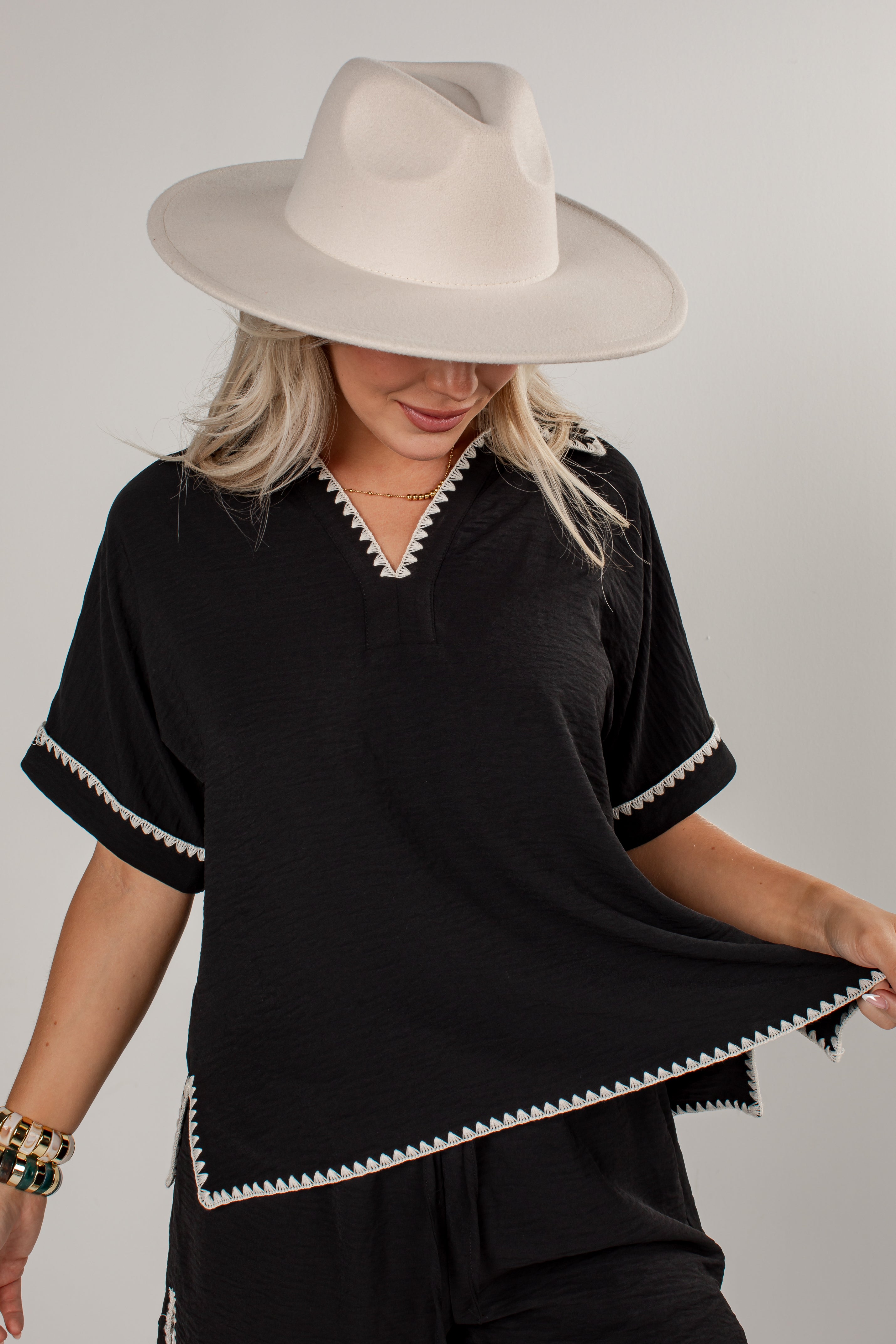 A woman wearing a black short-sleeve top with white decorative trim along the V-neck, sleeve cuffs, and hem, paired with a wide-brimmed cream hat against a light gray studio background.