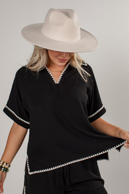 A woman wearing a black short-sleeve top with white decorative trim along the V-neck, sleeve cuffs, and hem, paired with a wide-brimmed cream hat against a light gray studio background.