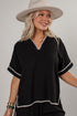 A woman wearing a black short-sleeve top with white embroidered trim, a wide-brim beige hat, and gold bracelets, photographed against a plain light gray studio background.