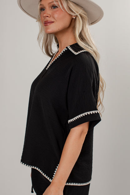 A woman wearing a black short-sleeve top with white zigzag trim along the collar, sleeve cuffs, and hem, paired with a wide-brimmed beige hat, photographed against a light gray studio background.