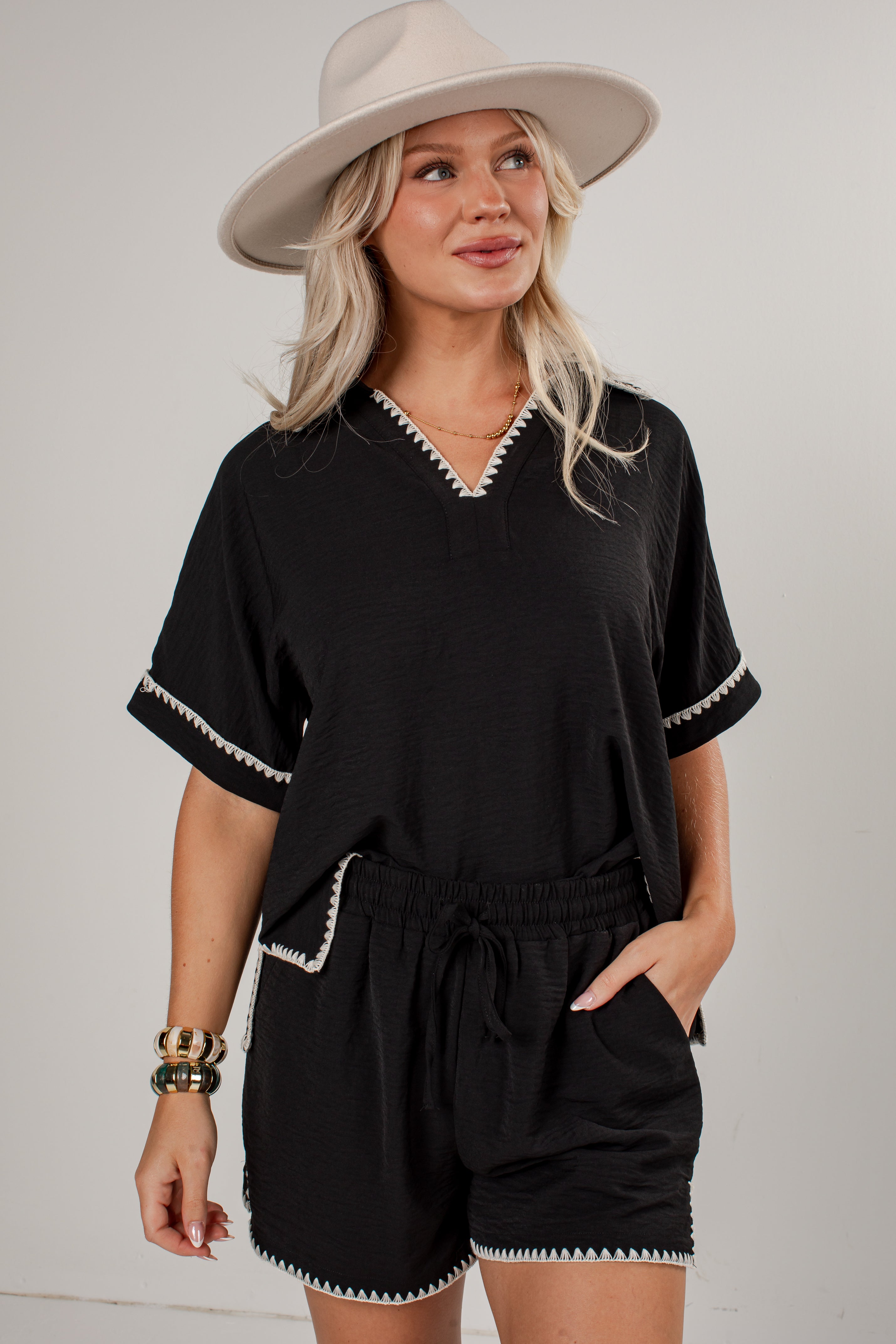 A woman modeling a black short-sleeve romper with white contrast stitching details, paired with a beige wide-brim hat, against a light gray studio background.