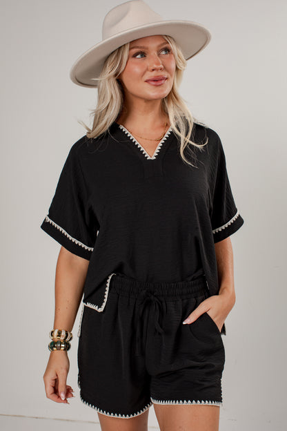 A woman modeling a black short-sleeve romper with white contrast stitching details, paired with a beige wide-brim hat, against a light gray studio background.
