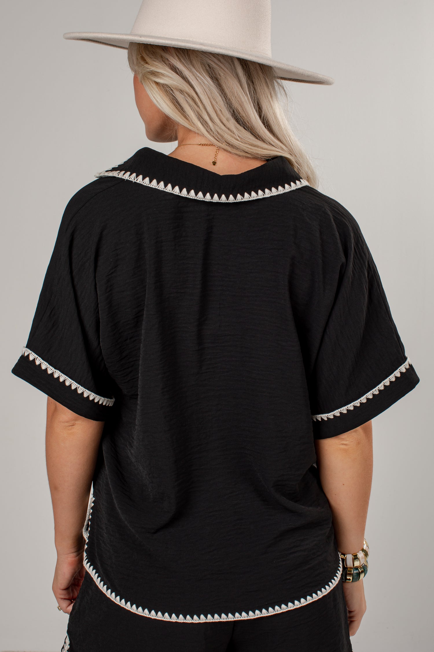 A woman viewed from the back wearing a black short-sleeve top with white geometric trim along the collar, sleeve cuffs, and hem, paired with a light beige wide-brim hat against a plain light gray studio background.