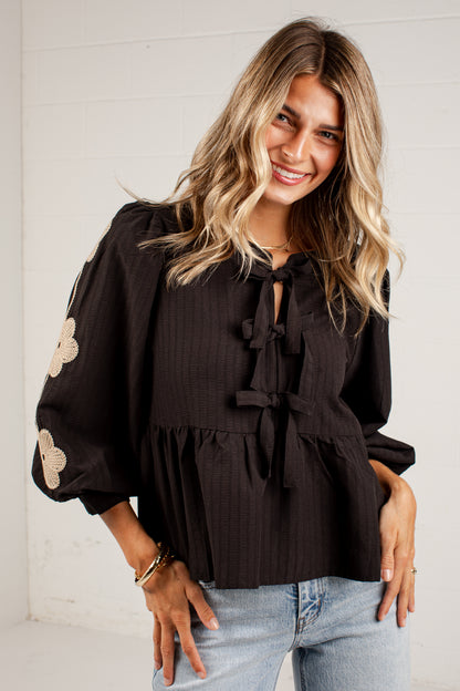 A woman wearing a black long-sleeve blouse with beige floral appliques on the sleeves and a tie-neck detail, paired with light blue jeans, standing against a white paneled wall.