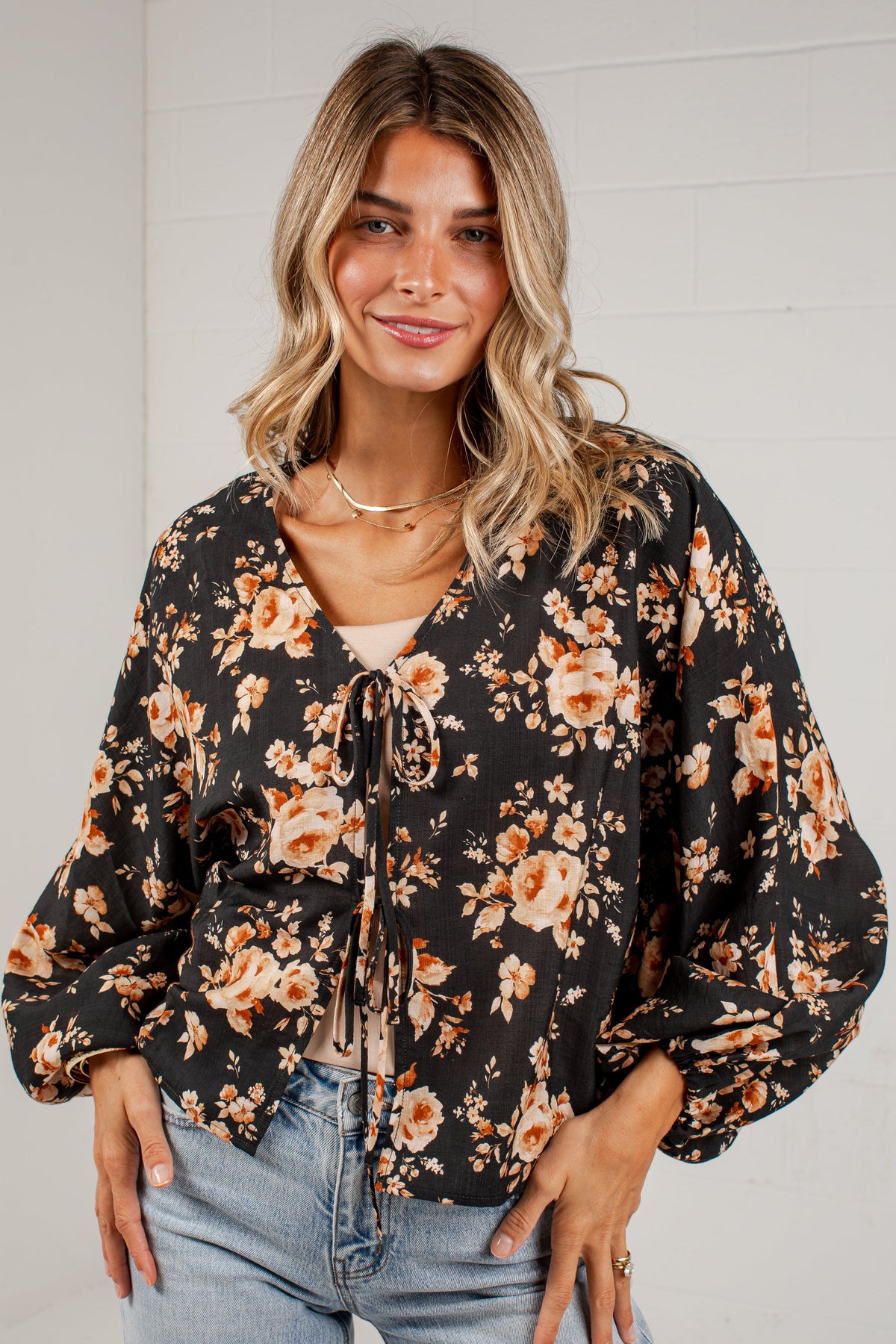 A woman wearing a black long-sleeve blouse with a warm-toned floral print and tie-front detail, paired with light-wash blue jeans, standing against a white paneled wall.