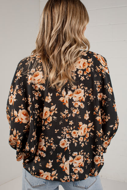 A woman viewed from behind wearing a black long-sleeve blouse with a warm-toned floral print in orange, beige, and cream, paired with light blue jeans against a white brick wall.