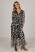 A woman modeling a long-sleeve maxi dress with a black, gray, and white floral pattern, paired with brown heeled ankle boots, against a plain light gray studio background.