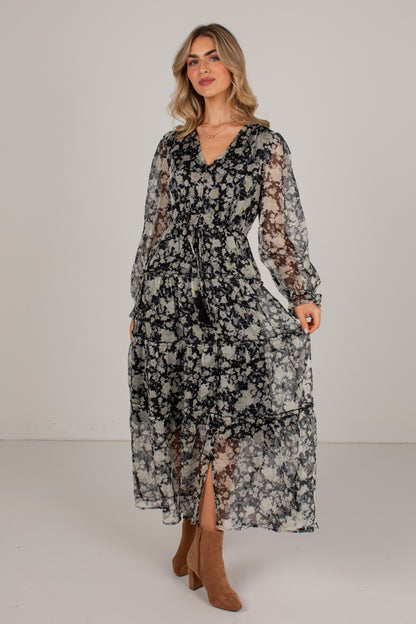 A woman modeling a long-sleeve midi dress with a black and beige floral pattern, sheer lace sleeves, and a V-neckline, paired with brown suede boots against a plain studio background.