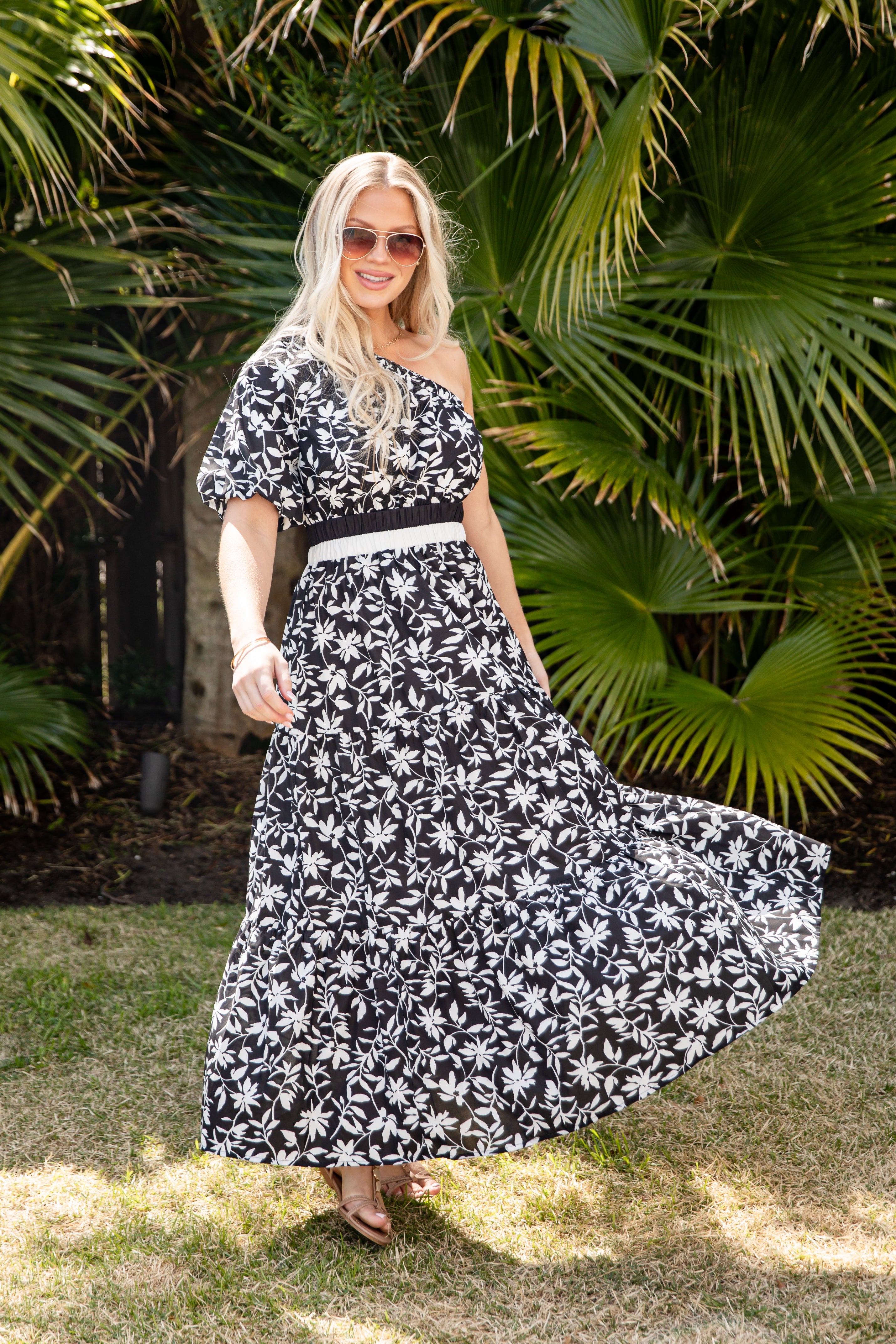 Woman in a black and white floral dress standing in front of green foliage.