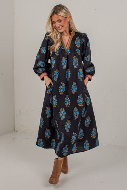 A woman modeling a long-sleeved black maxi dress with a blue and gold floral pattern, paired with brown suede boots, in a studio setting.