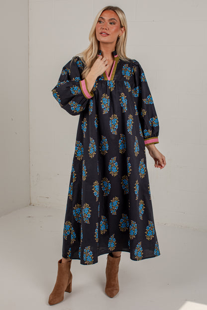 A woman modeling a long-sleeved black maxi dress with a blue and gold floral pattern, pink trim, and brown ankle boots, standing in a white studio corner.