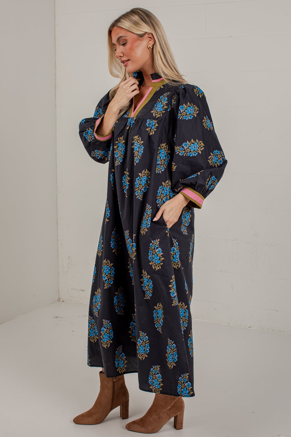 A woman modeling a long-sleeved navy maxi dress with a blue, gold, and pink floral pattern, paired with brown ankle boots, in a white studio setting.