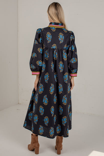 A woman viewed from behind wearing a long black midi dress with a colorful blue, gold, and pink floral pattern, paired with brown ankle boots in a white studio setting.
