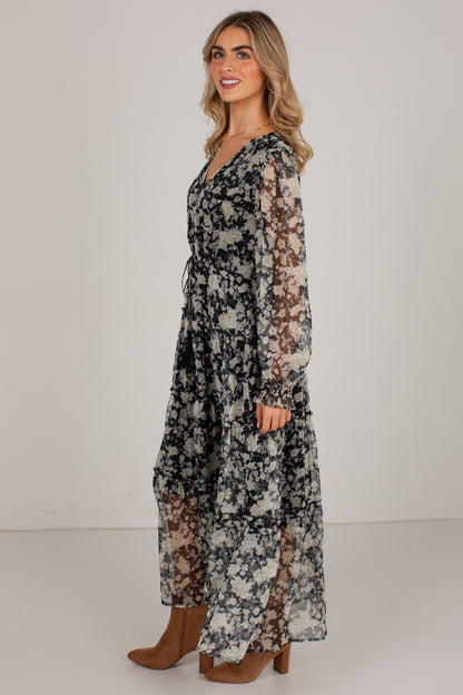 A woman modeling a long-sleeve maxi dress with a black, white, and gray floral pattern and sheer mesh panels, paired with brown ankle boots, against a plain light gray studio background.