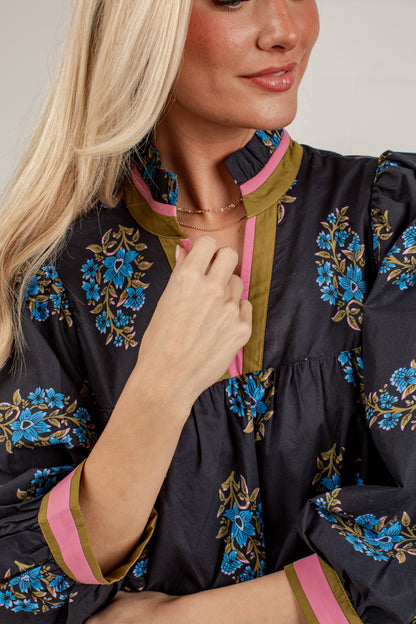 A close-up of a woman wearing a dark navy floral blouse with blue, gold, and pink accents. The blouse features a ruffled high collar, a gathered waist, and long sleeves with pink and olive green trim.