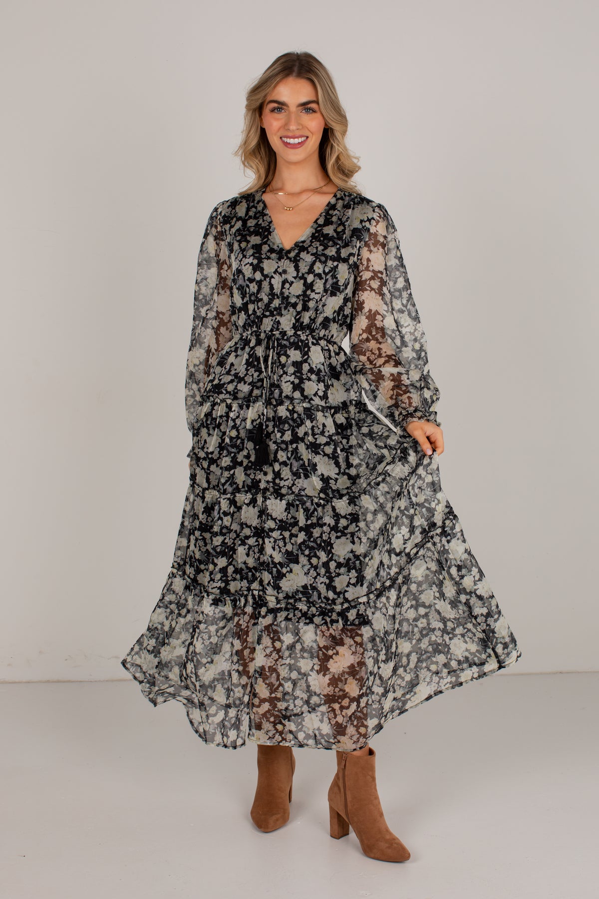 A woman modeling a long-sleeved, tiered maxi dress with a black and white floral pattern, paired with brown suede ankle boots, against a plain light gray studio background.