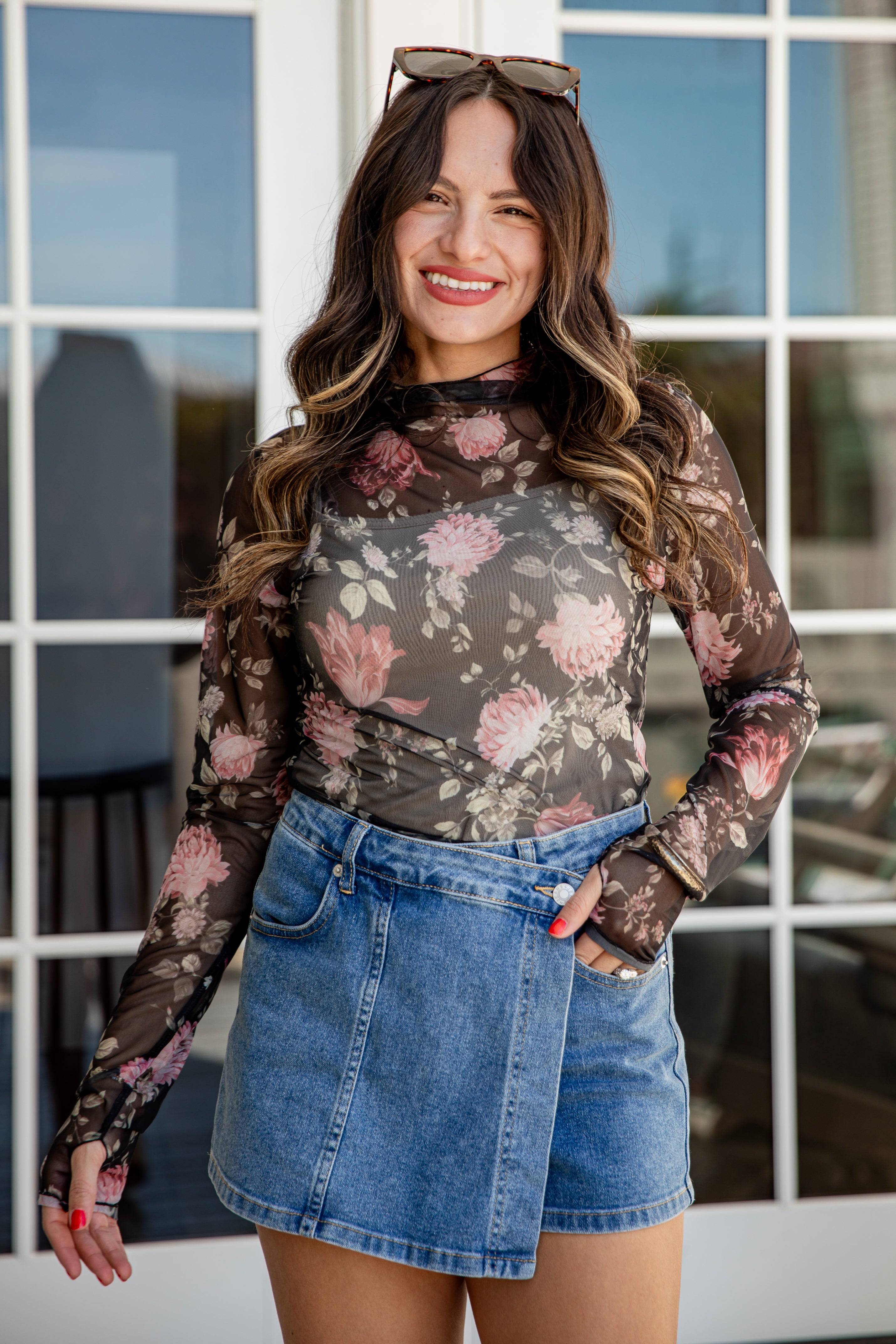 Woman wearing a floral long-sleeve top and denim skirt standing in front of a window.