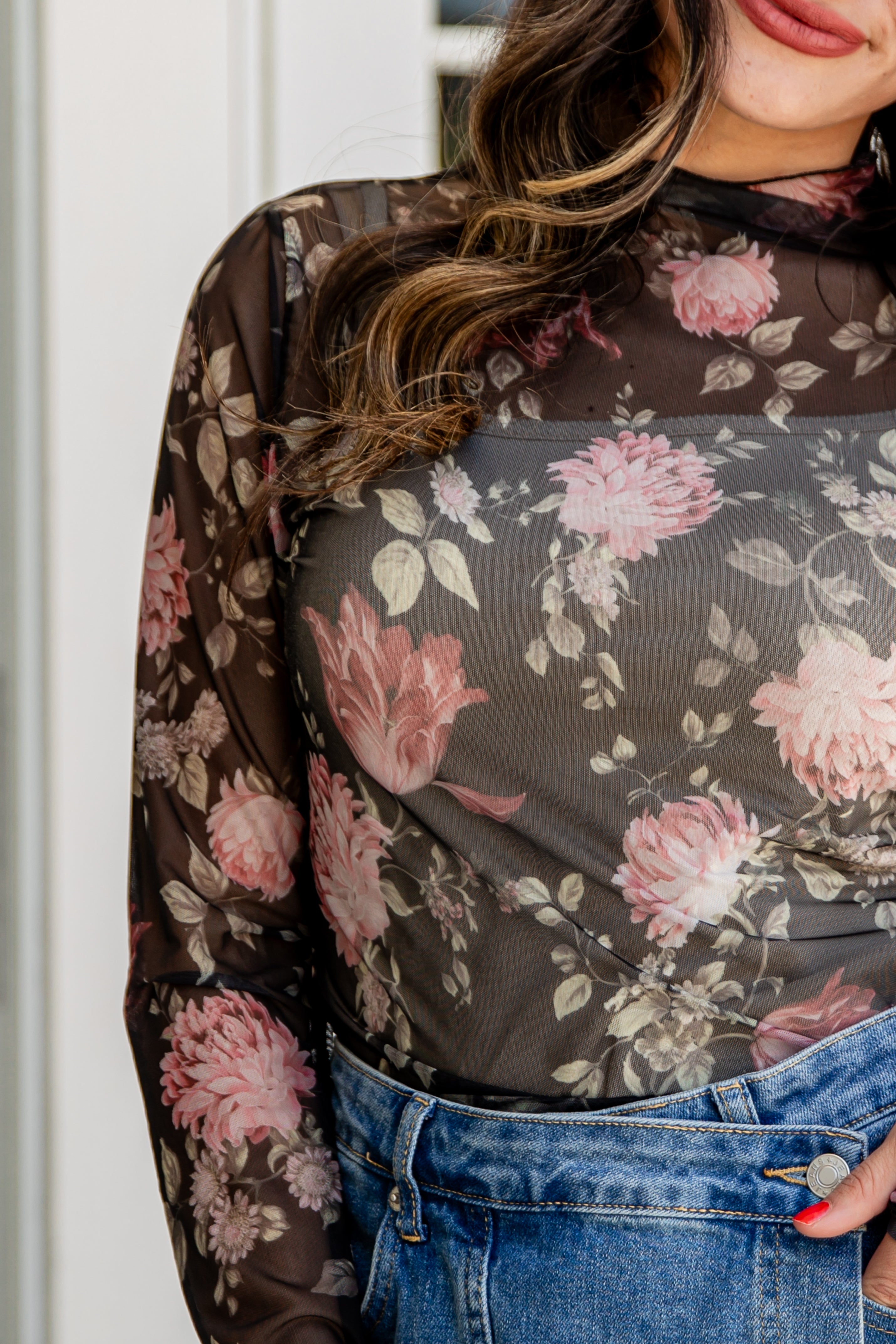 Person wearing a floral sheer top with pink flowers on a dark background, paired with blue jeans.