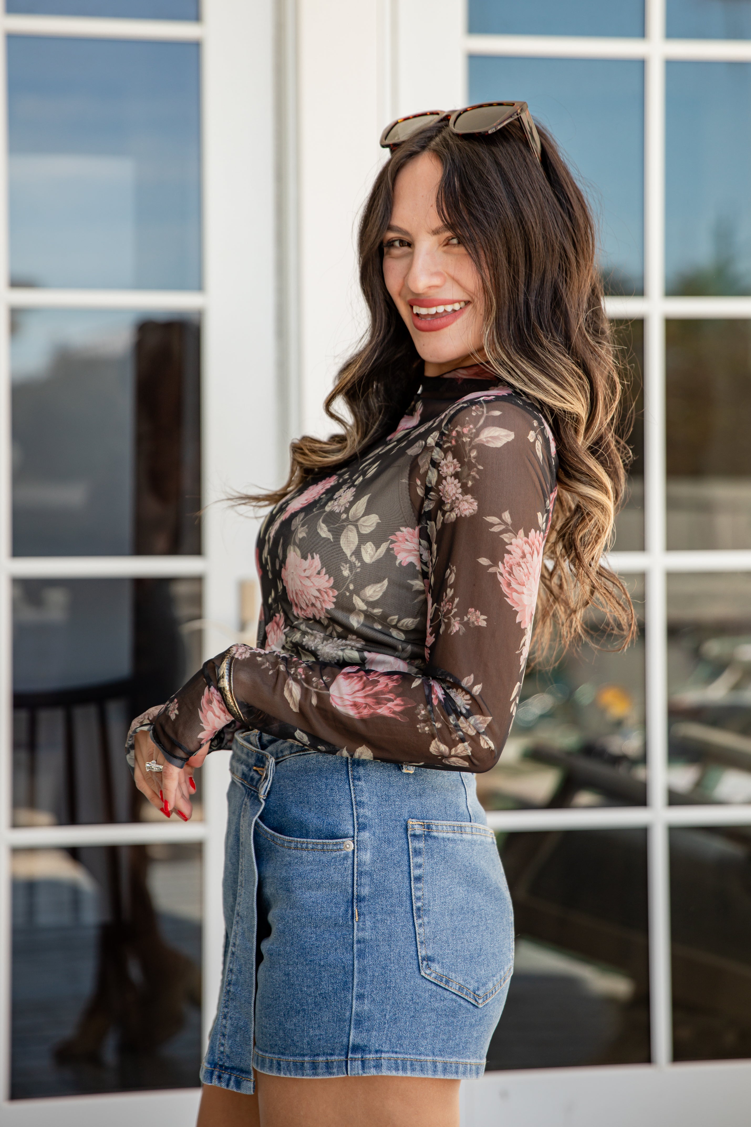 Woman wearing a floral long-sleeve top and denim shorts standing in front of a glass door.