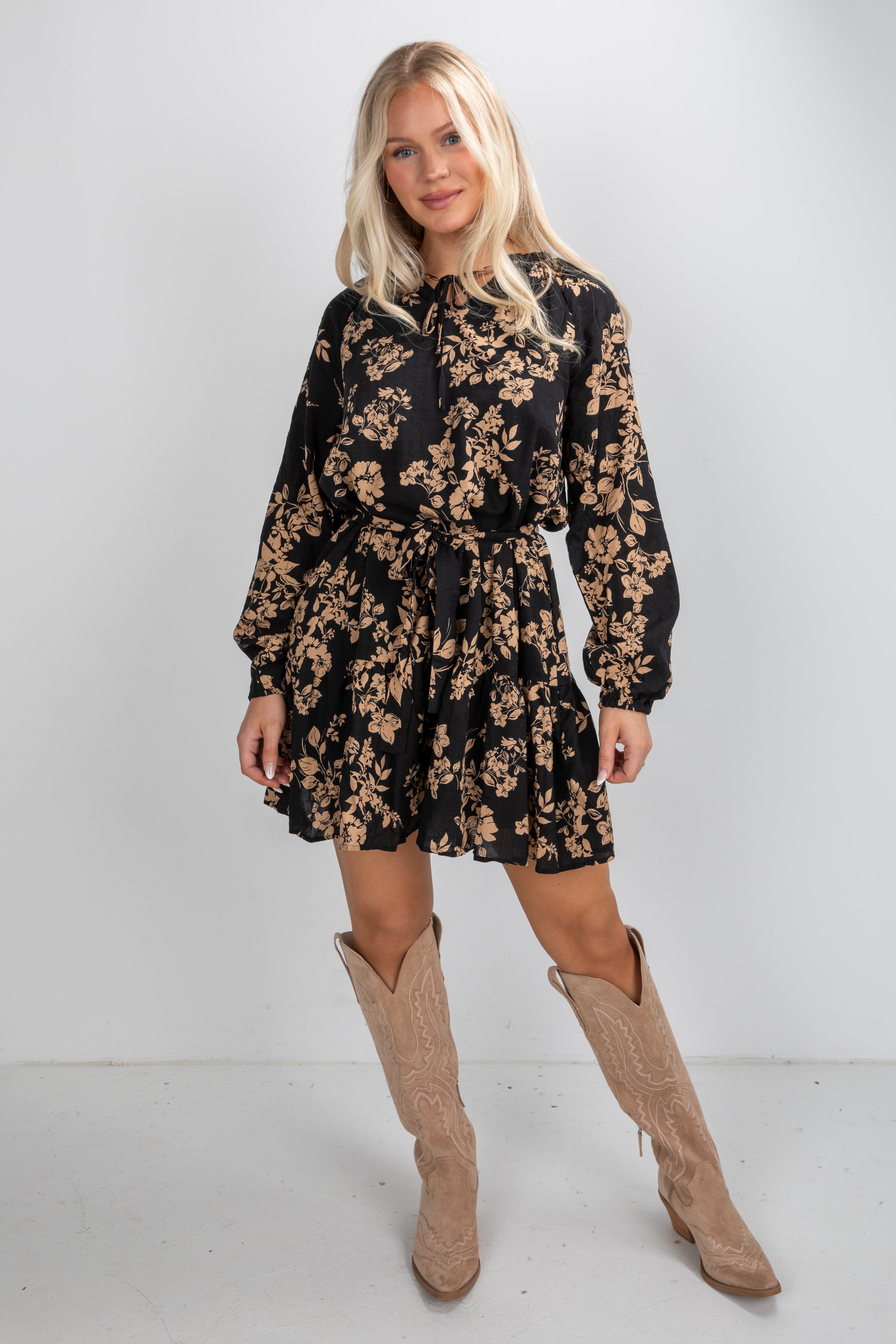 Woman wearing a black floral dress with long sleeves and beige knee-high boots on a white background