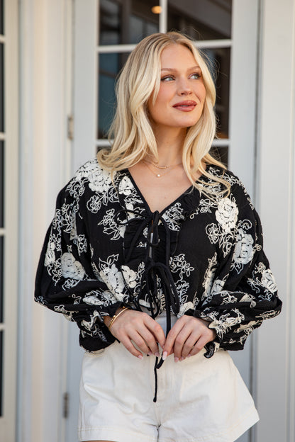 Woman wearing a black floral blouse and white pants standing in front of a glass door.
