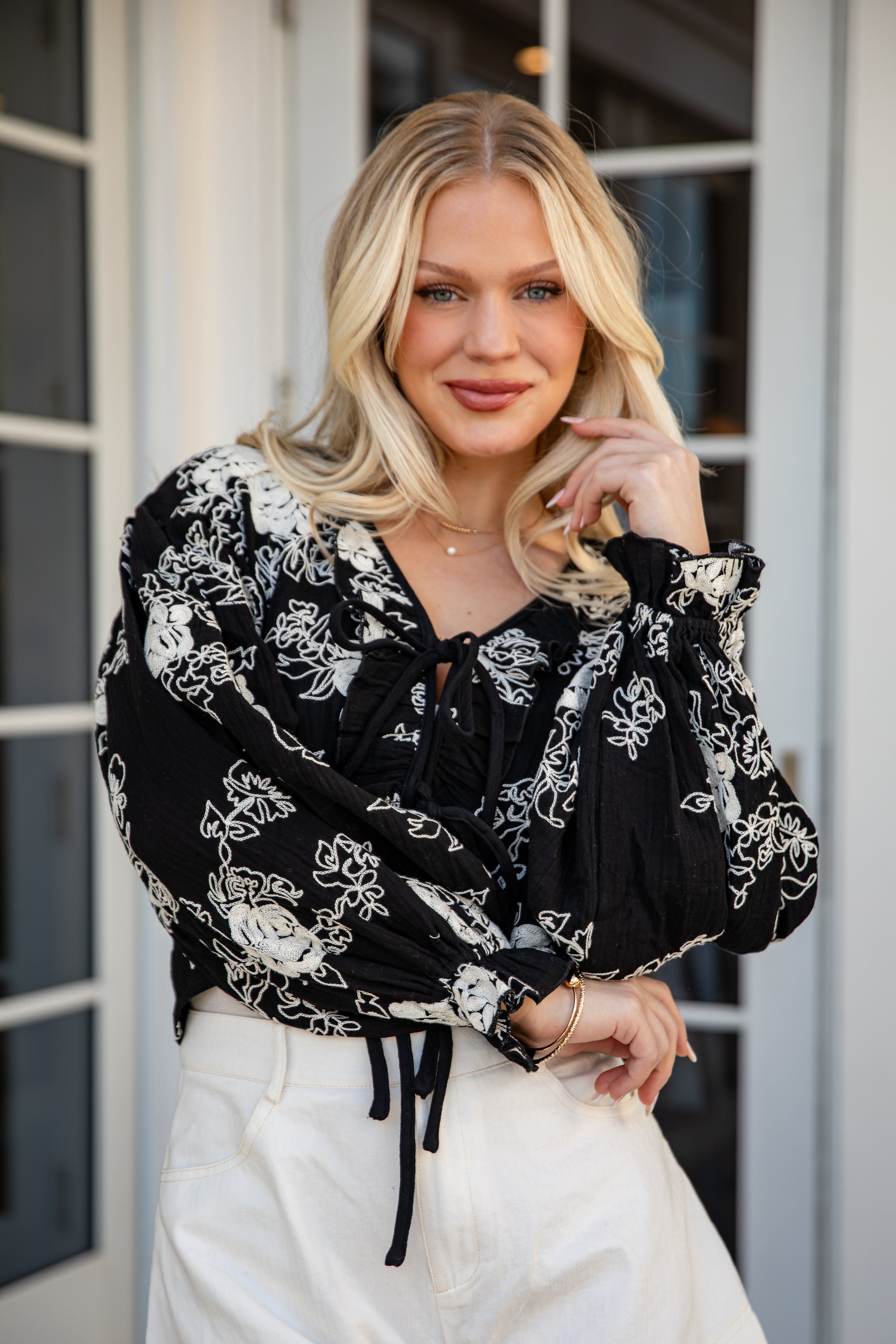 Woman wearing a black floral blouse and white pants standing in front of a glass door.