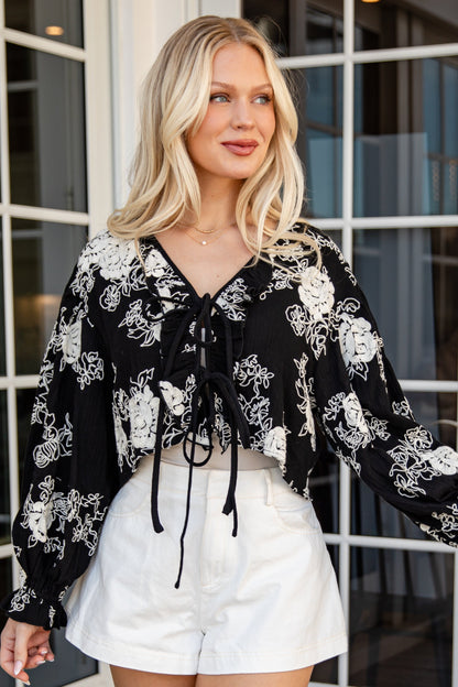 Woman wearing a black floral blouse and white shorts standing in front of glass doors.