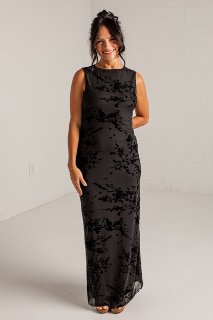 A woman modeling a sleeveless black maxi dress with a tonal floral lace overlay, standing against a white studio wall.