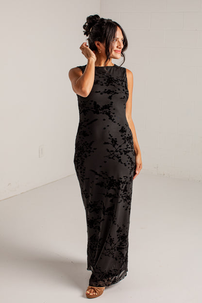 A woman modeling a sleeveless black maxi dress with a tonal floral lace overlay, standing in a bright white studio.