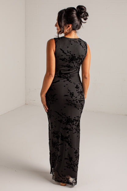 A woman viewed from behind wearing a sleeveless black maxi dress with a tonal floral lace overlay, standing in a white studio corner.