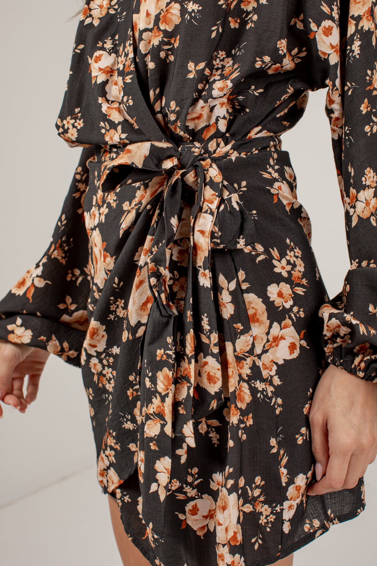 A close-up of a model wearing a black long-sleeve mini dress with a warm-toned floral print in orange, beige, and cream, featuring a tie waist belt.