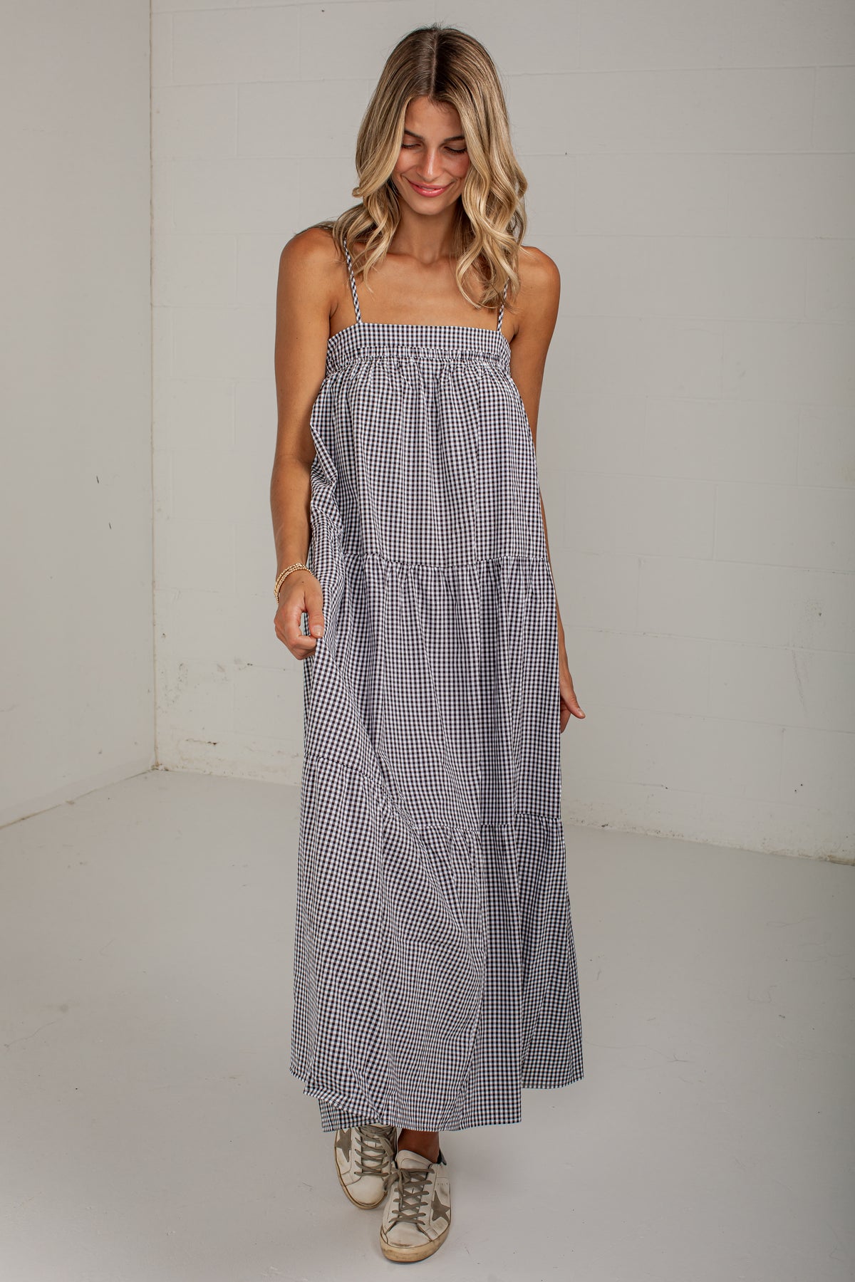 A woman modeling a sleeveless, tiered maxi dress with a small black and white gingham pattern, standing against a plain white studio wall.