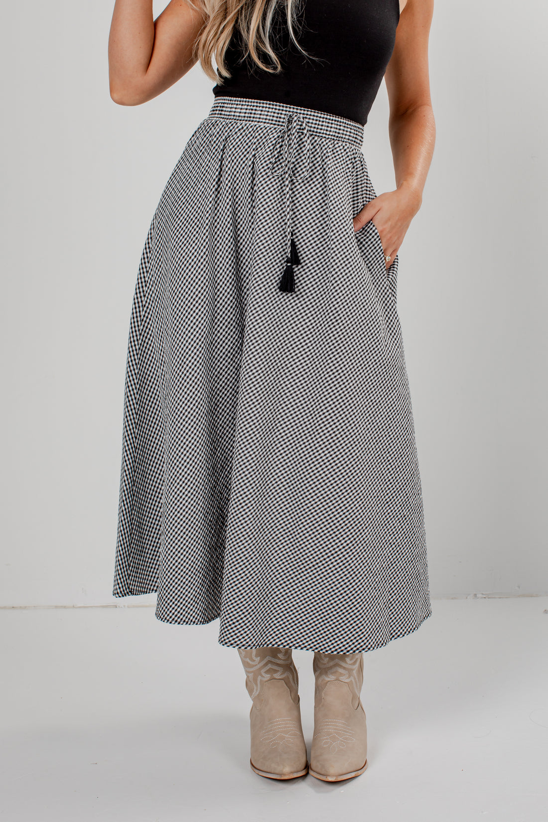 A woman wearing a high-waisted, A-line midi skirt with a black and white micro-pattern, a black top, and beige cowboy boots, standing against a light gray studio background.