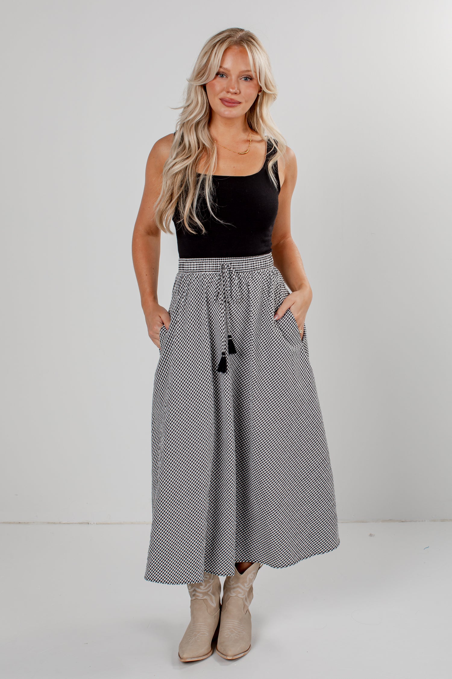 A woman modeling a high-waisted, maxi-length skirt with a black and white micro-check pattern, a drawstring waist, and black tassels, paired with a black sleeveless top and beige boots.